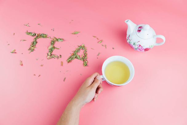 dámská ruka drží šálek čaje s bílou konvicí na boku a slova čaj napsaný s volným listovým čajem na jasně růžovém pozadí - Fotografie, Obrázek
