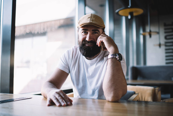 Portrait of smiling caucasian mature male in casual wear and hat sitting at coffee shop enjoying free time, happy prosperous bearded man owner of small business looking at camera satisfied with job - Photo, Image
