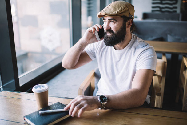 Casual cheerful man making phone call while sitting at wooden table in cafe having notepad with pen and disposable cup of coffee near - Foto, afbeelding