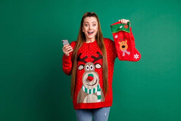 Photo of attractive excited lady hold newyear stocking x-mas eve presents telephone shopping internet buy gifts family tradition wear red ugly pullover isolated green color background - Foto, imagen