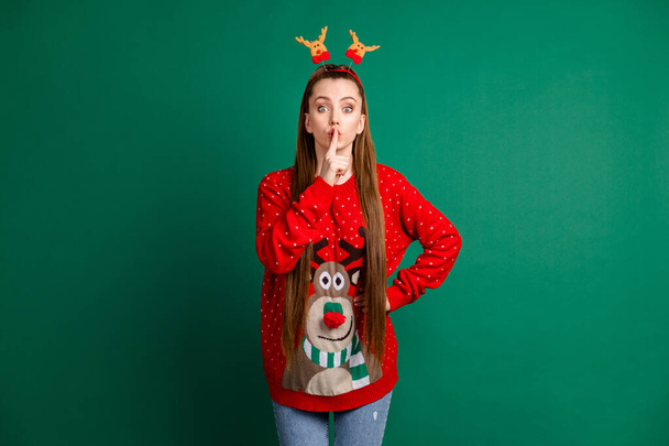 Photo of attractive cute lady long brown tail finger on lips ask friends keep silence boyfriend x-mas surprise wear deer horns headwear red ornament pullover isolated green color background - Foto, Imagen