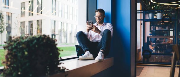 Positive young businessman in casual clothes and eyeglasses sitting near window in modern workspace and reading message on smartphone while resting during work - Fotó, kép