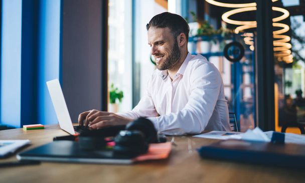 Side view of cheerful bearded male entrepreneur in formal shirt sitting at wooden desk in modern workspace and working on project on laptop - Photo, Image