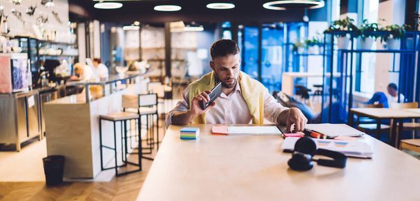 Современный бородатый мужчина в светлой рубашке говорит по телефону и работает с документами, сидя в уютном хорошо освещенном кафе - Фото, изображение