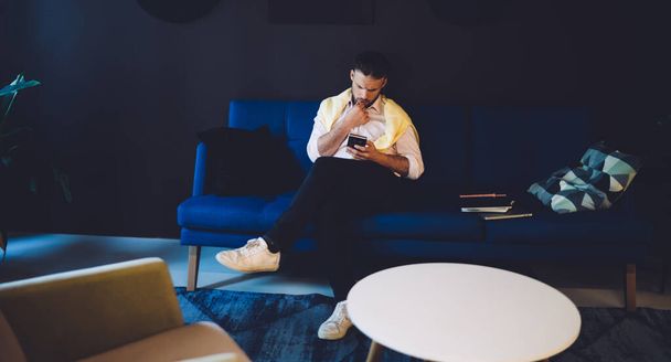 Seriózní muž podnikatel ve stylovém ležérní oblečení sedí na pohovce v moderním pracovním prostoru a pomocí smartphone při odpočinku po práci na projektu - Fotografie, Obrázek