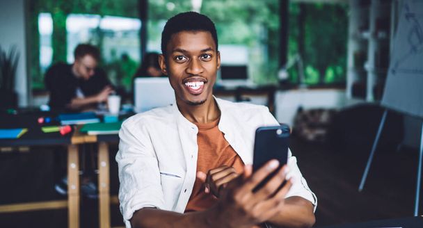 Portrait of cheerful male blogger holding modern cellphone gadget and smiling at camera during free time for online web networking, toothy African American millennial connecting to 4g on smartphone - Fotoğraf, Görsel