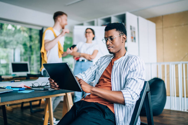 Serious African American spectacled man in casual clothes surfing tablet in university while cheerful male and female students interacting behind - Foto, Imagen