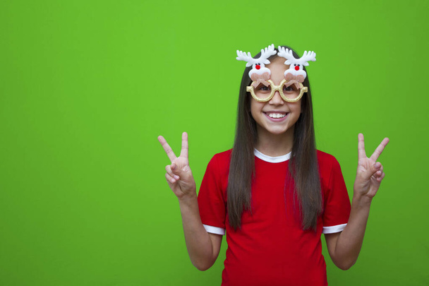 Χαμογελαστό κορίτσι με σκούρα μαλλιά σε κόκκινο μπλουζάκι και με αστεία γυαλιά, για τα Χριστούγεννα και την Πρωτοχρονιά δείχνει δύο δάχτυλα μέχρι την ειρήνη - Φωτογραφία, εικόνα