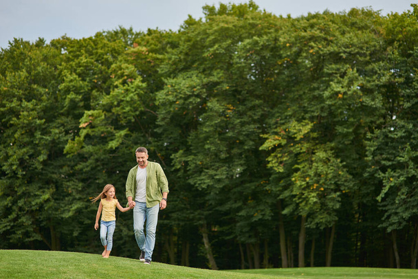 Aufnahme in voller Länge, in der sich der junge Vater und seine kleine Tochter an einem Sommertag beim Spazierengehen im schönen grünen Park an den Händen halten - Foto, Bild