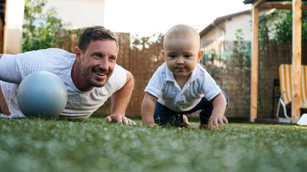 Ένα χαριτωμένο μωρό και ο χαμογελαστός πατέρας του παίζουν σε ένα χορτάρι με μια μπάλα - Φωτογραφία, εικόνα