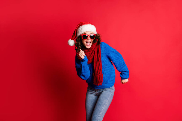 Készen állok felgyújtani a táncparkettet. Fénykép portré izgatott lány ünnepli téli ünnepek visel szemüveg sál santa kalap kék pulóver elszigetelt élénk piros színű háttér - Fotó, kép