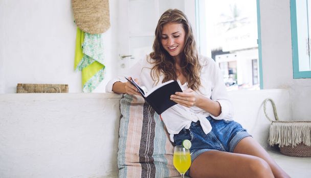 Captivated woman in denim shorts reading book while sitting on couch with glass of refreshing drink in sunny modern apartment - 写真・画像