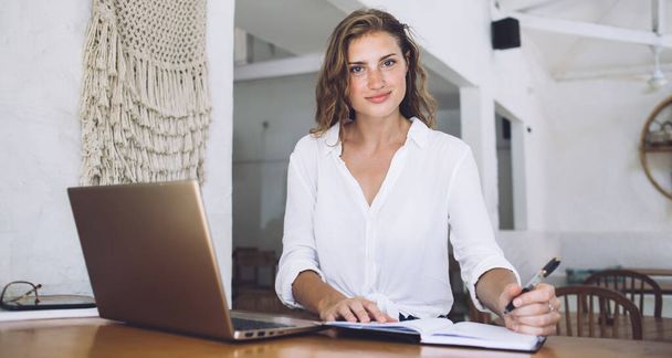 Smiling adult business lady holding pen and looking at camera while sitting alone at table with notebook and laptop in cafe - Foto, immagini