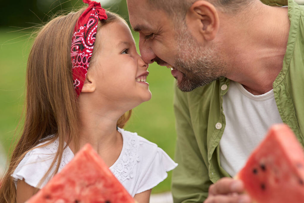 close-up van schattig klein meisje en haar liefhebbende vader aanraken neuzen tijdens het eten van watermeloen, familie met een picknick in het groene park op een zomerse dag - Foto, afbeelding