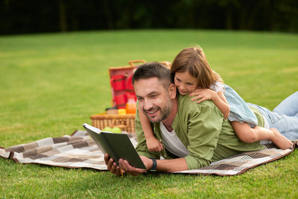 Curious little girl distracting, playing with her dad when he reads a book while lying on a blanket. Happy father and his daughter having a picnic in the park - Φωτογραφία, εικόνα