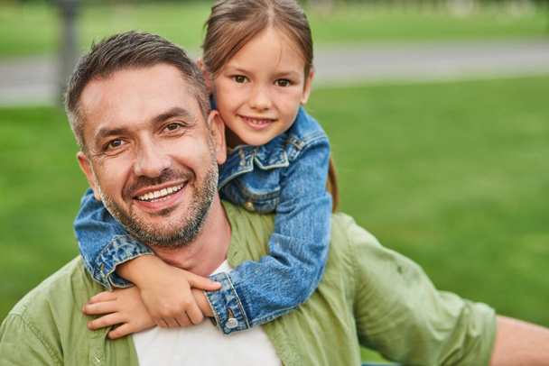 Portrait of young joyful father and his cute little daughter looking at camera and hugging while spending time together outdoors, sitting on a green grass - Valokuva, kuva
