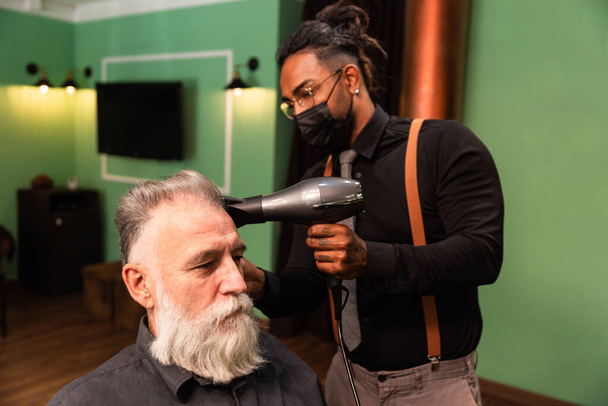 Jonge Afro-Amerikaanse getatoeëerde kapper kamt en föhns het haar van een volwassen blanke man met een witte baard - Foto, afbeelding