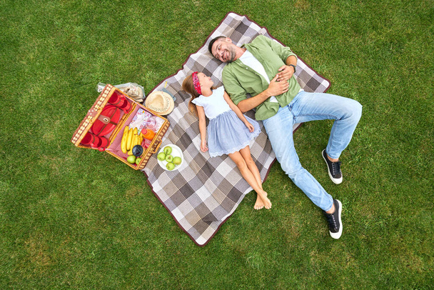 A boldog apa és bájos kislánya egymásra mosolyognak, a takarón fekszenek, miközben piknikeznek a zöld parkban. - Fotó, kép