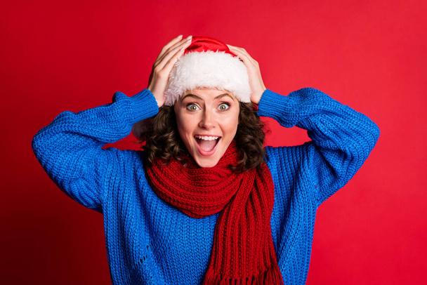 Photo portrait of young girl excited for unbelievable winter sales discounts open mouth holding head with hands wearing blue pullover scarf santa hat isolated on bright red colored background - 写真・画像