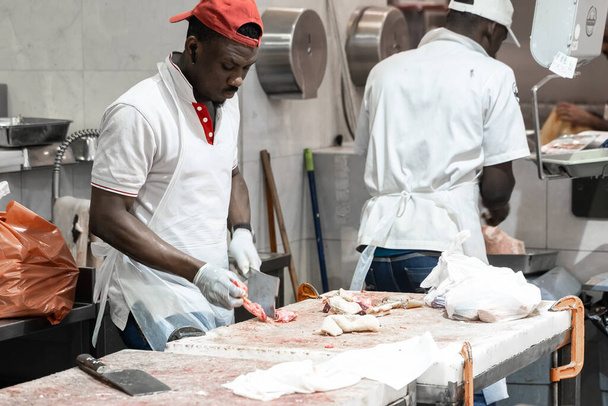 DUBAI, UAE - MARCH 2, 2020: African american butcher boning fresh ham in a modern butcher shop with metal safety mesh glove. A man cook cuts meat with a knife in a restaurant. - Φωτογραφία, εικόνα
