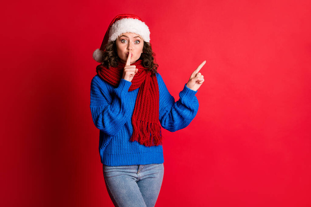 Photo portrait festive girl tells you keep quiet about surprise gift pointing at blank space wearing blue pullover scarf santa hat isolated on bright red colored background - Fotoğraf, Görsel