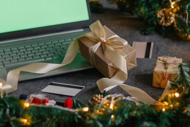 Christmas shopping online with a credit card for the holiday. Laptop with gifts on the table next to fir branches and a garland of lights. - Photo, Image