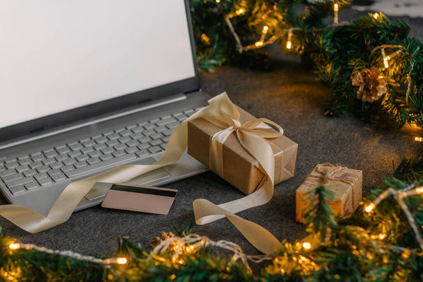 Christmas shopping online with a credit card for the holiday. Laptop with gifts on the table next to fir branches and a garland of lights. - 写真・画像