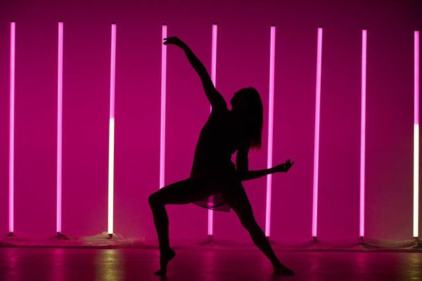 Silhouette of a young woman dancer dancing and posing in a room with neon lamps. A slender modern model in a beautiful pose on a purple background. - Fotó, kép