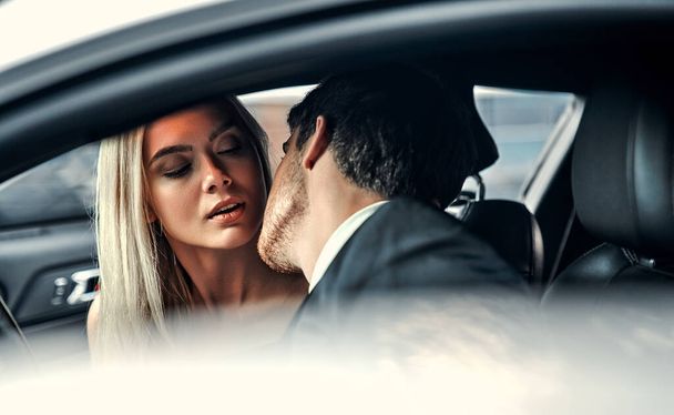 Passionate couple making love in car. Attractive blonde in elegant black dress and handsome businessman in suit are kissing in car. - Zdjęcie, obraz