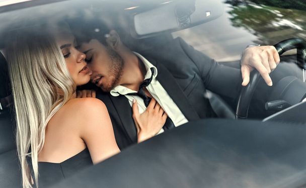 Passionate couple making love in car. Attractive blonde in elegant black dress and handsome businessman in suit are kissing in car. - Foto, Bild