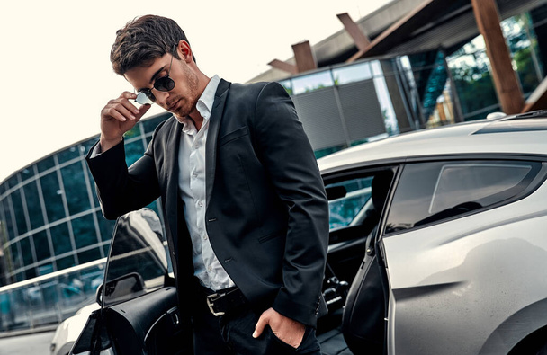 Handsome young businessman in suit is standing near his sport car in front of modern business center - 写真・画像