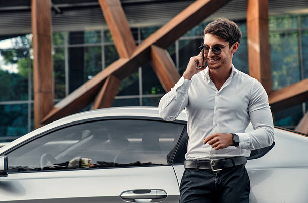 Guapo joven hombre de negocios en traje está de pie cerca de su coche deportivo en frente de un moderno centro de negocios y hablando por teléfono móvil - Foto, imagen