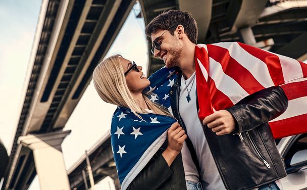 Beautiful young couple in casual clothes near sport car with American flag in hands. - Valokuva, kuva