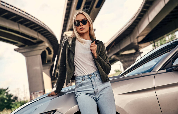 Attractive young blonde in casual clothes  and sunglasses standing near sport car on the street. Urban style. - Fotografie, Obrázek