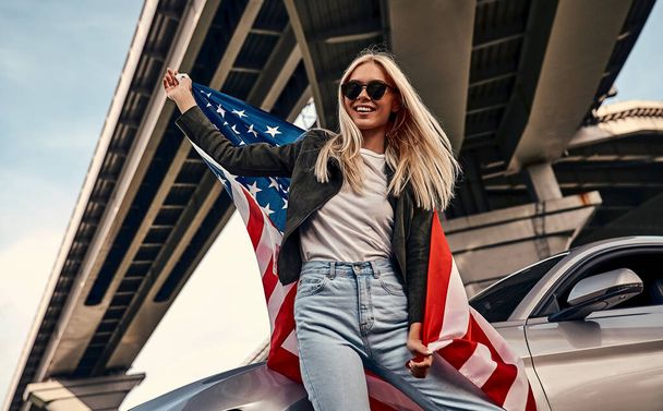 Attractive young blonde in casual clothes  and sunglasses standing near sport car on the street with American flag in hands.  Urban style. - Фото, зображення