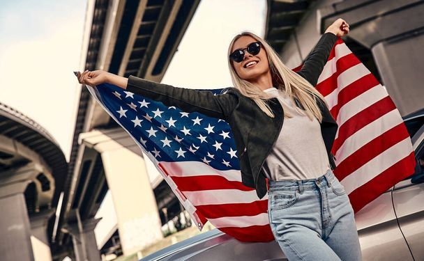 Ελκυστική νεαρή ξανθιά σε casual ρούχα και γυαλιά ηλίου στέκεται κοντά σπορ αυτοκίνητο στο δρόμο με αμερικανική σημαία στα χέρια. Αστικό στυλ. - Φωτογραφία, εικόνα