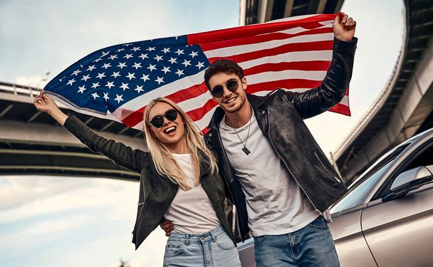 Beautiful young couple in casual clothes near sport car with American flag in hands. - Foto, imagen