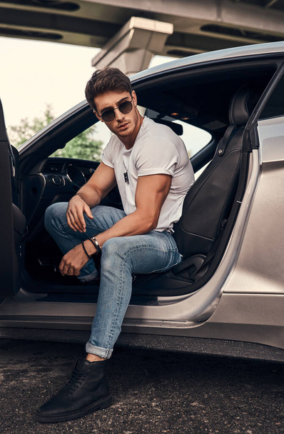 Bonito jovem de óculos de sol e roupas casuais sentado em seu carro esporte moderno - Foto, Imagem