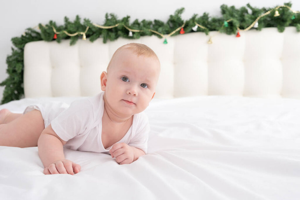 ξανθό αγοράκι ξαπλωμένο σε λευκό κρεβάτι στο σπίτι με χριστουγεννιάτικα στολίδια. - Φωτογραφία, εικόνα