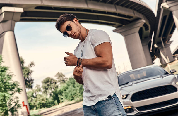 Handsome young man in casual clothes standing near his modern sport car on the street - 写真・画像