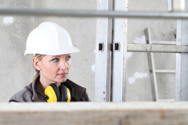donna operaio edile ritratto indossando casco bianco e cuffie di protezione dell'udito, tenendo una scala sul cantiere interno sfondo con impalcatura - Foto, immagini