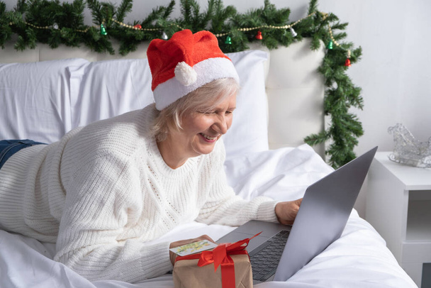 Femme âgée positive avec smartphone faisant du shopping pour Noël à la maison au lit. concept de vieillesse. - Photo, image