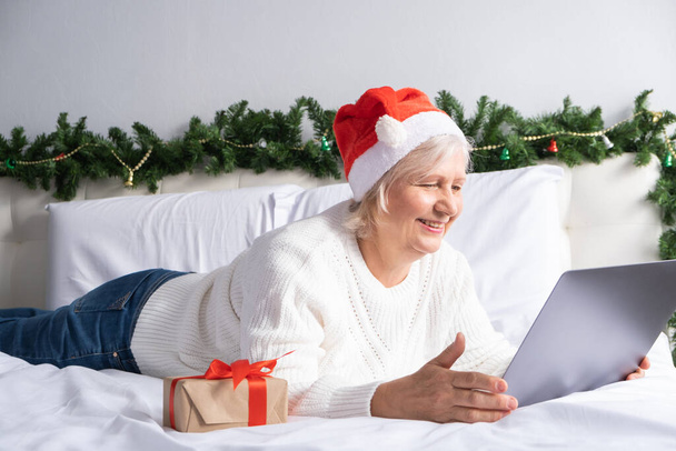 Karácsonyi online üdvözlet. boldog idős nők télapó kalap otthon ágyban laptop. - Fotó, kép