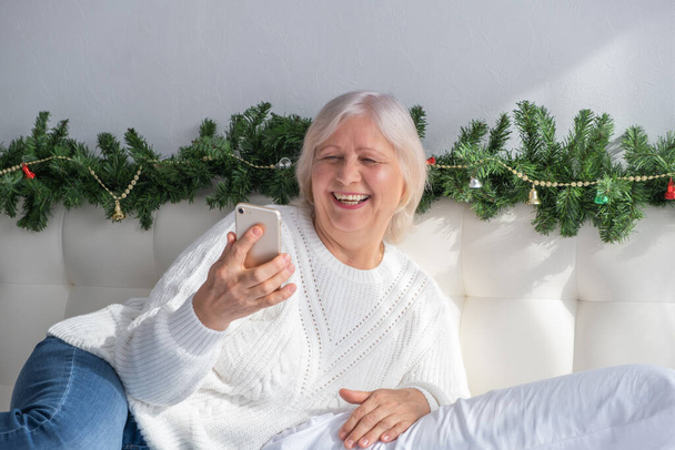 Positive senior female with smartphone doing shopping for Christmas at home in bed. old age concept. - Foto, immagini