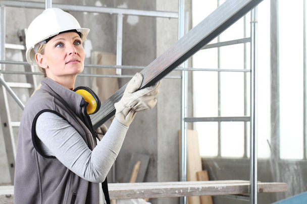 woman construction worker builder portrait wearing white helmet and hearing protection headphones, holding a metal stud for drywall on interior site building background with scaffolding - Photo, Image