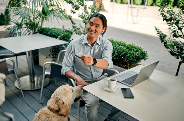 Asian businessman working in the city with laptop wearing wireless earphones. Confident young man sitting in cafe with dog. - Foto, Imagen