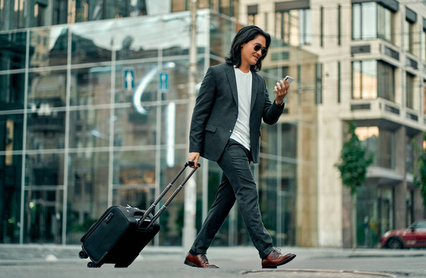 Asian businessman in the city. Confident young man walking outdoor in suit and sunglasses with smart phone and suitcase in hands - Foto, Bild