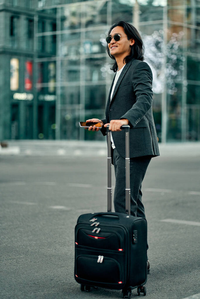 Asian businessman in the city. Confident young man standing outdoor in suit and sunglasses with smart phone and suitcase in hands - Fotoğraf, Görsel