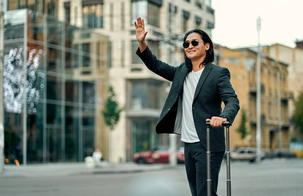 Asian businessman in the city. Confident young man standing outdoor in suit and sunglasses with suitcase in hands and trying to stop taxi - Foto, Imagem