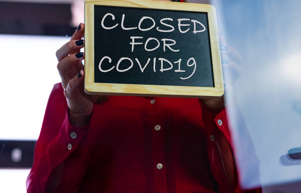 Vista dal basso di una donna caucasica con una camicetta rossa che mette un segno di chiuso da merluzzo bianco19 sulla porta di vetro della sua attività. - Foto, immagini